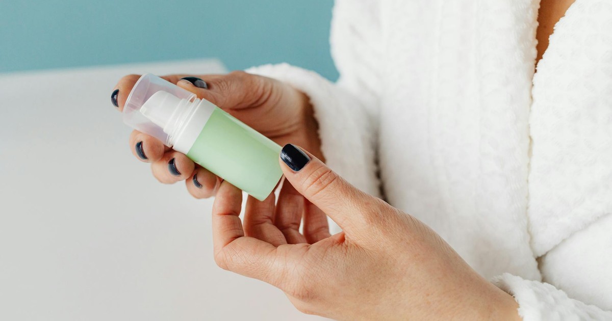 A person in a bathrobe holding a skincare product, focusing on hands and bottle.