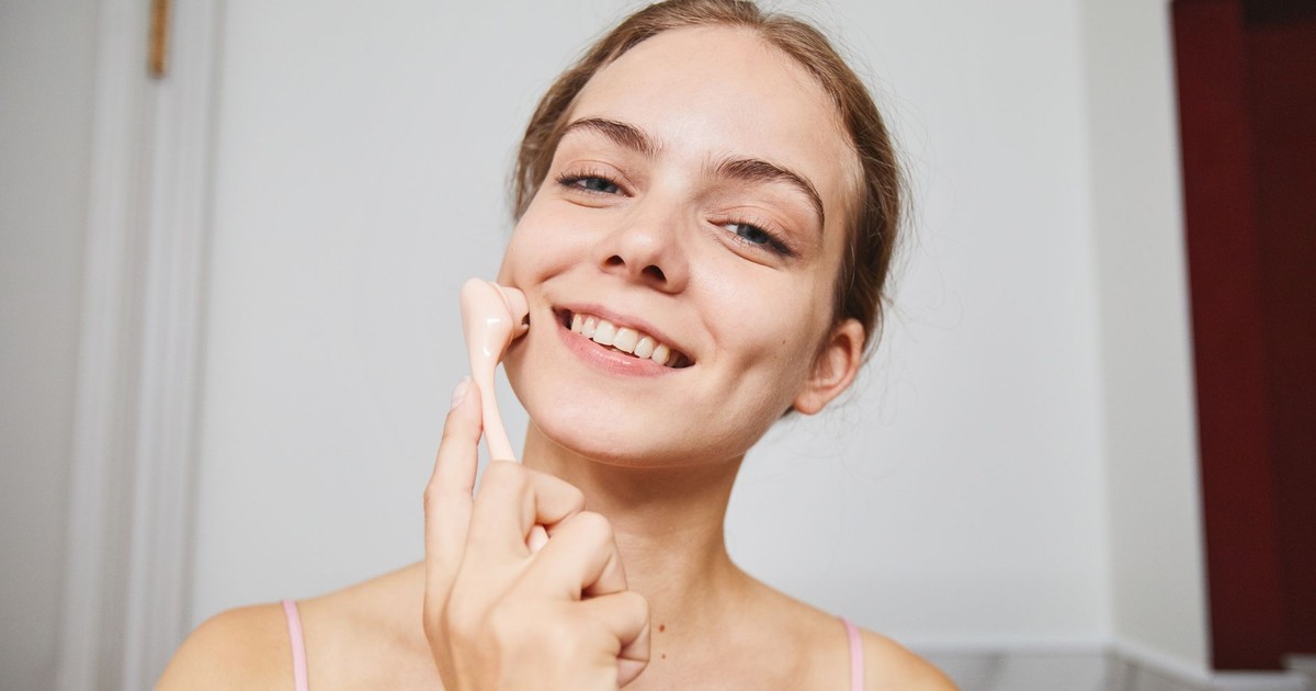 Happy woman enjoying a skincare routine using a facial massage roller indoors.