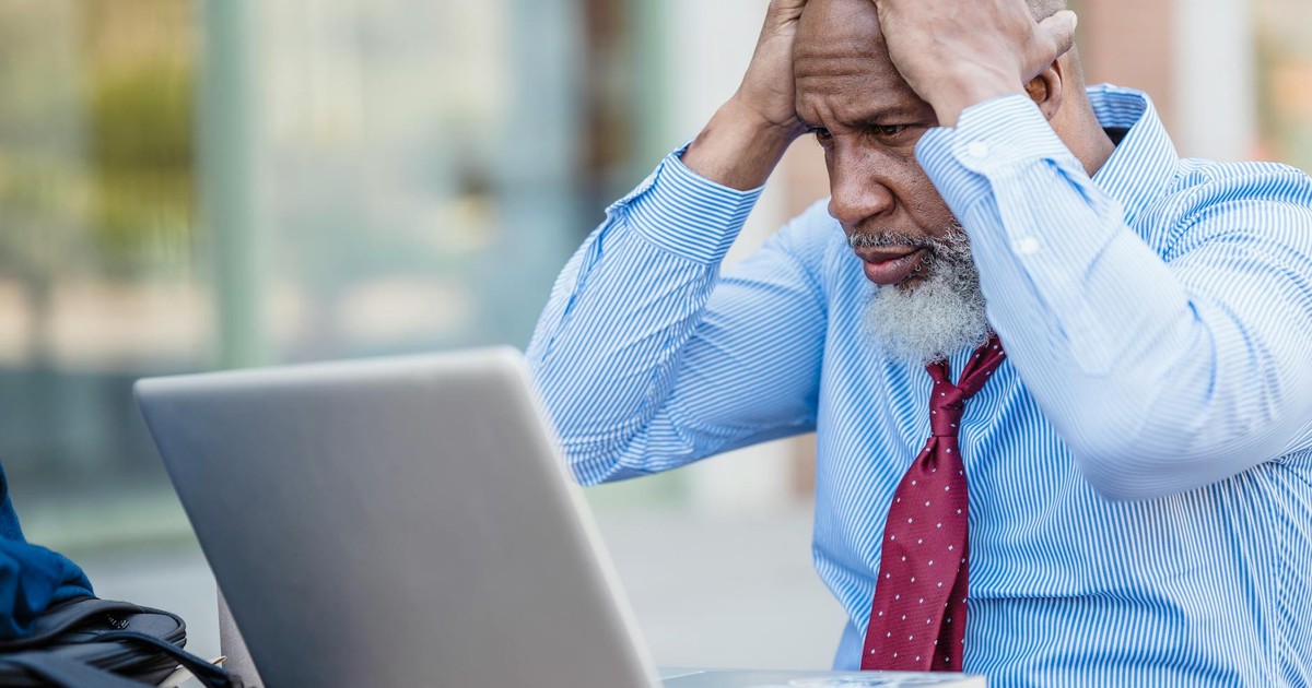 Anxious senior African American male entrepreneur in formal classy shirt and tie sitting at table with laptop and grabbing head
