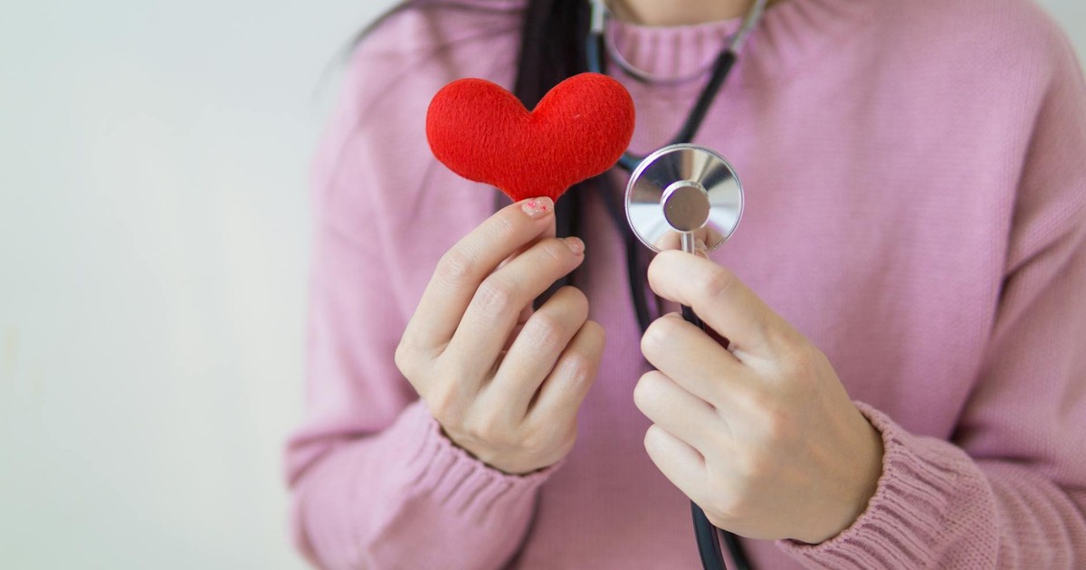 Unrecognizable female in pink sweater with stethoscope on neck standing on white background with red heart in hand in daylight