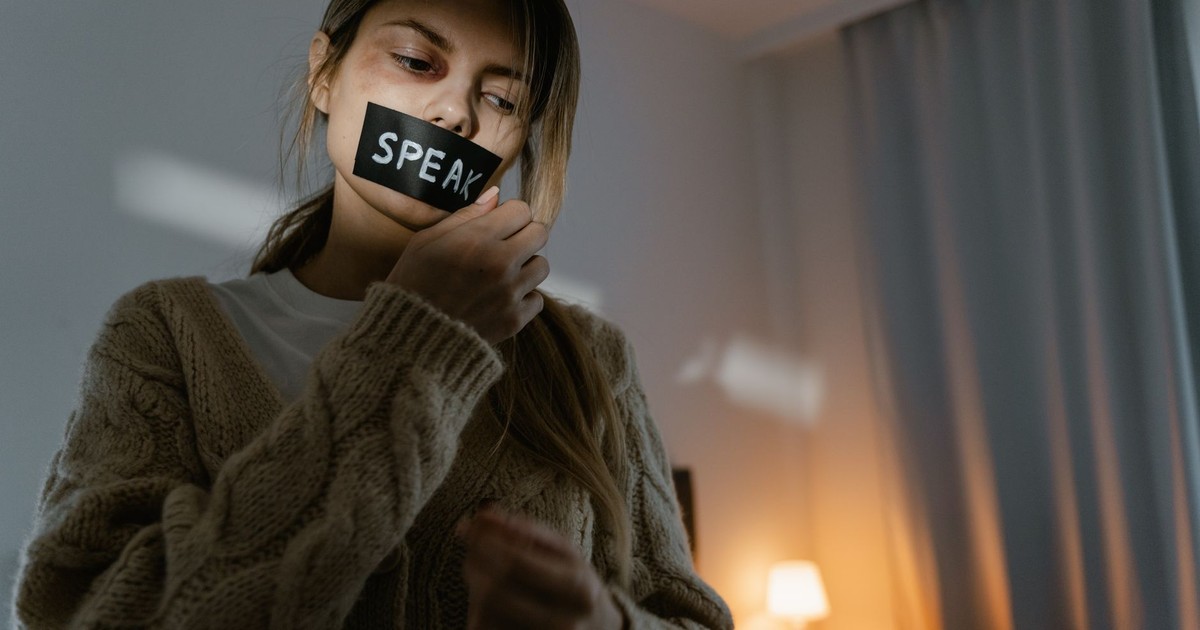 A woman standing in a dimly lit room removing tape with 'SPEAK' written, symbolizing empowerment.