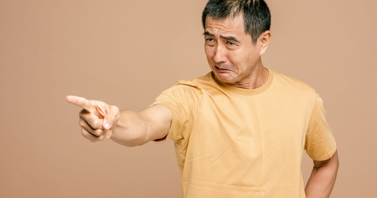 An adult man with a frustrated expression points his finger in anger against a brown background.