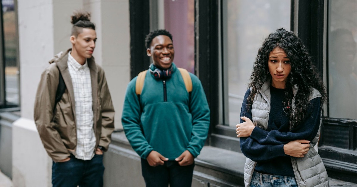 Cheerful multiracial young male students bullying sad frightened ethnic female groupmate standing on street with crossed arms