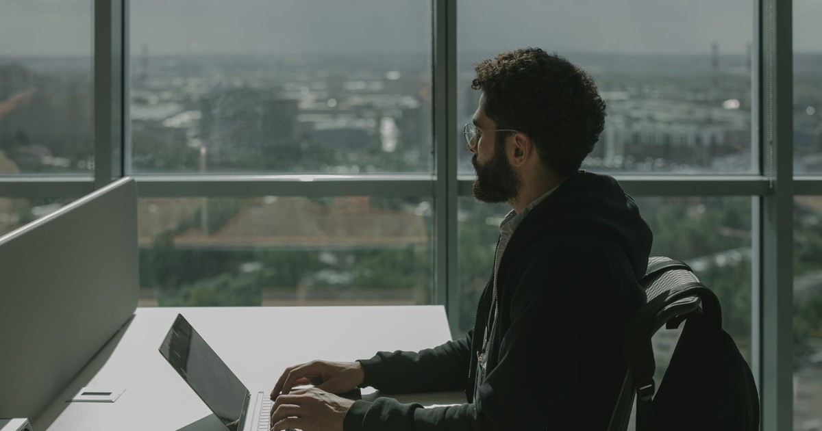 A man with facial hair types on a laptop at a modern office desk with a city view.