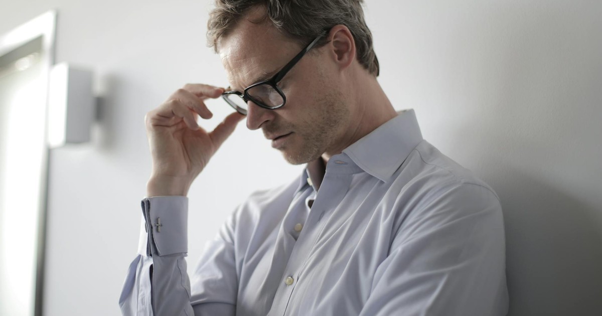 An adult man in a shirt and glasses appears stressed while leaning against a wall indoors.