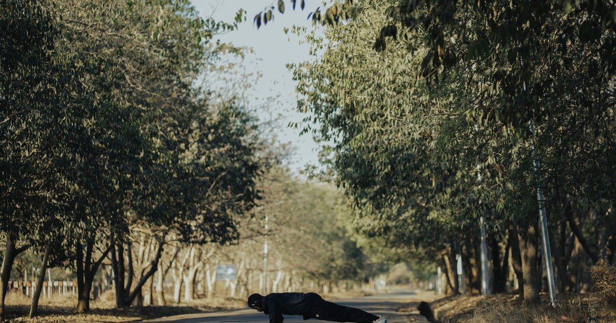 A man performs push-ups outdoors on a tree-lined pathway under the sun.