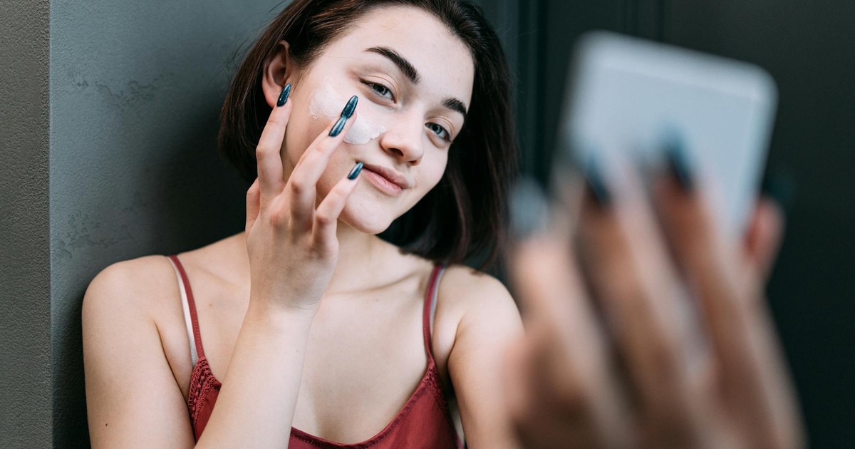 Young Caucasian woman applying moisturizer while smiling in front of a mirror indoors.