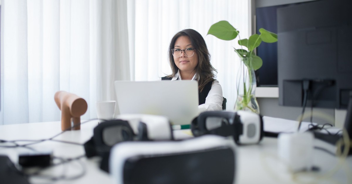 Asian woman in office using laptop with VR headsets on table, symbolizing modern workspace.