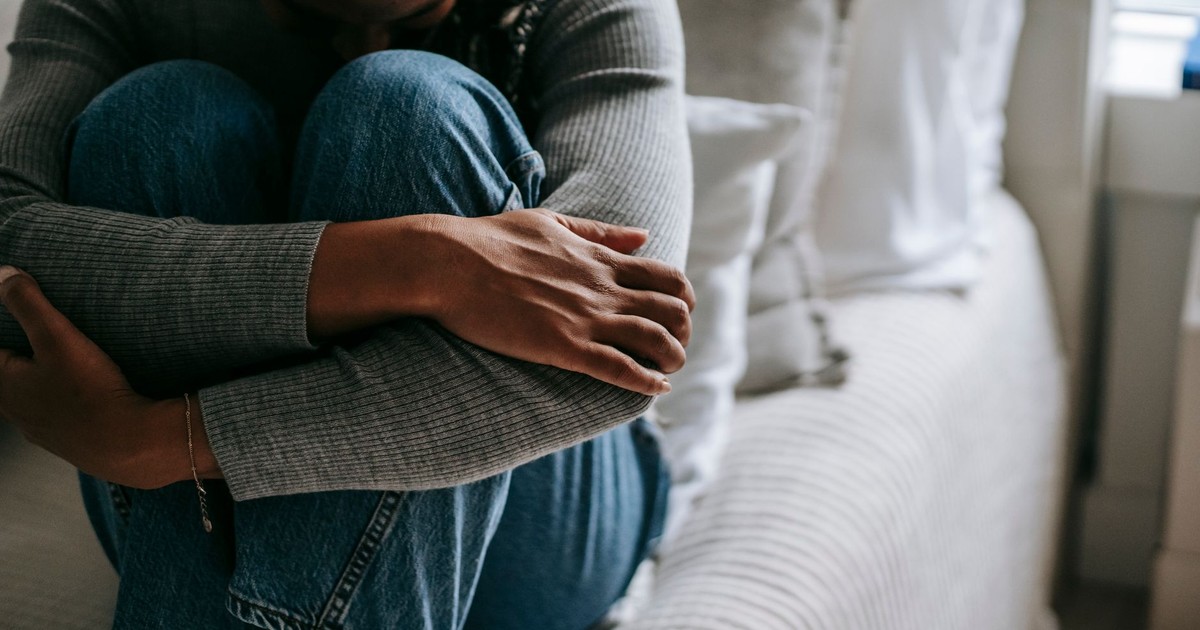 Crop unhappy mournful African American female wearing casual outfit embracing knees on bed and hiding face in sad thoughts