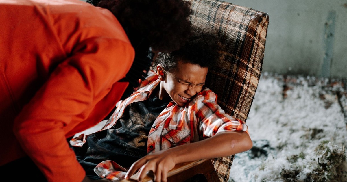 Child in distress on checkered chair with adult leaning over ominously inside a dimly lit room.