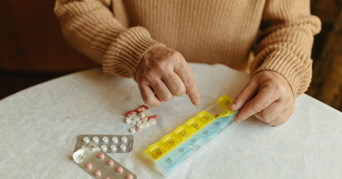 Senior adult organizing daily medications using a weekly pill organizer on a table indoors.