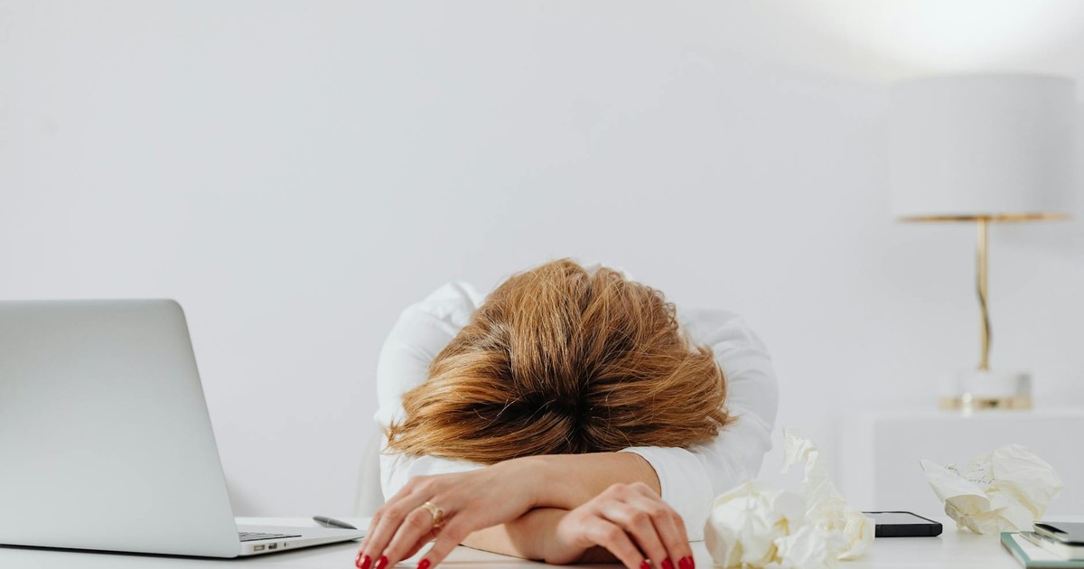 Tired woman resting head on arms at desk with laptop, showing fatigue in modern office.