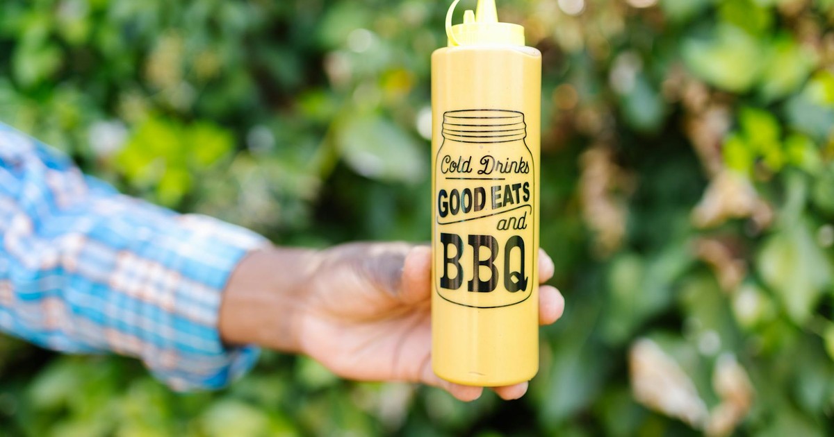 A close-up of a hand holding a yellow BBQ squeeze bottle with green foliage in the background.