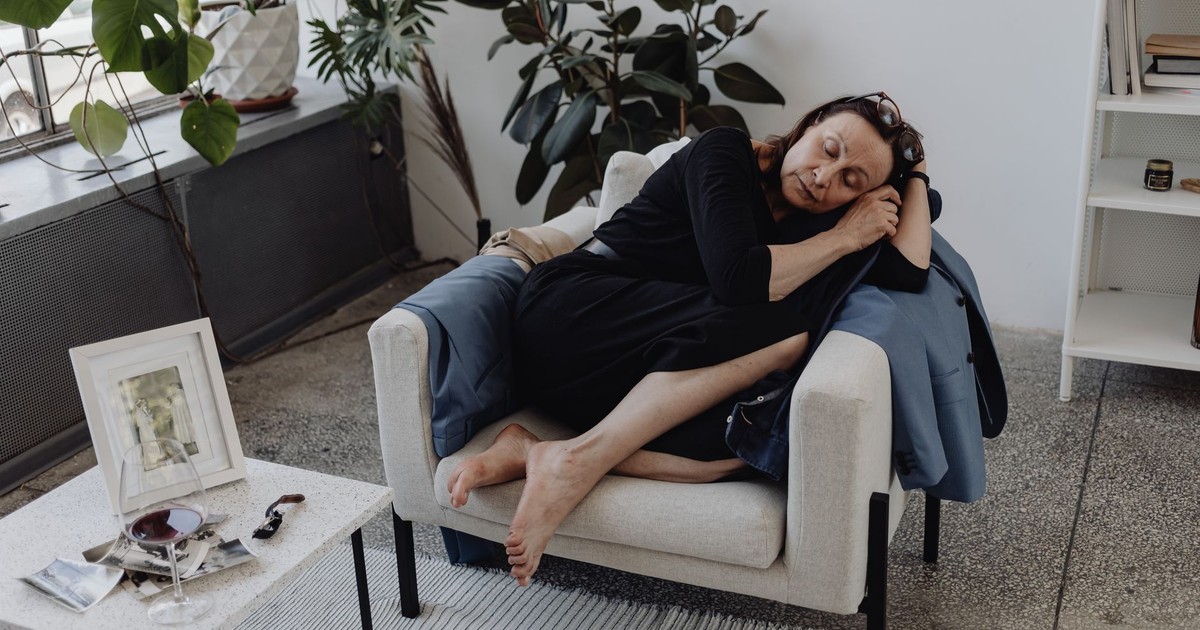 A senior woman asleep on an armchair in a cozy indoor setting, surrounded by plants and a table with a glass.