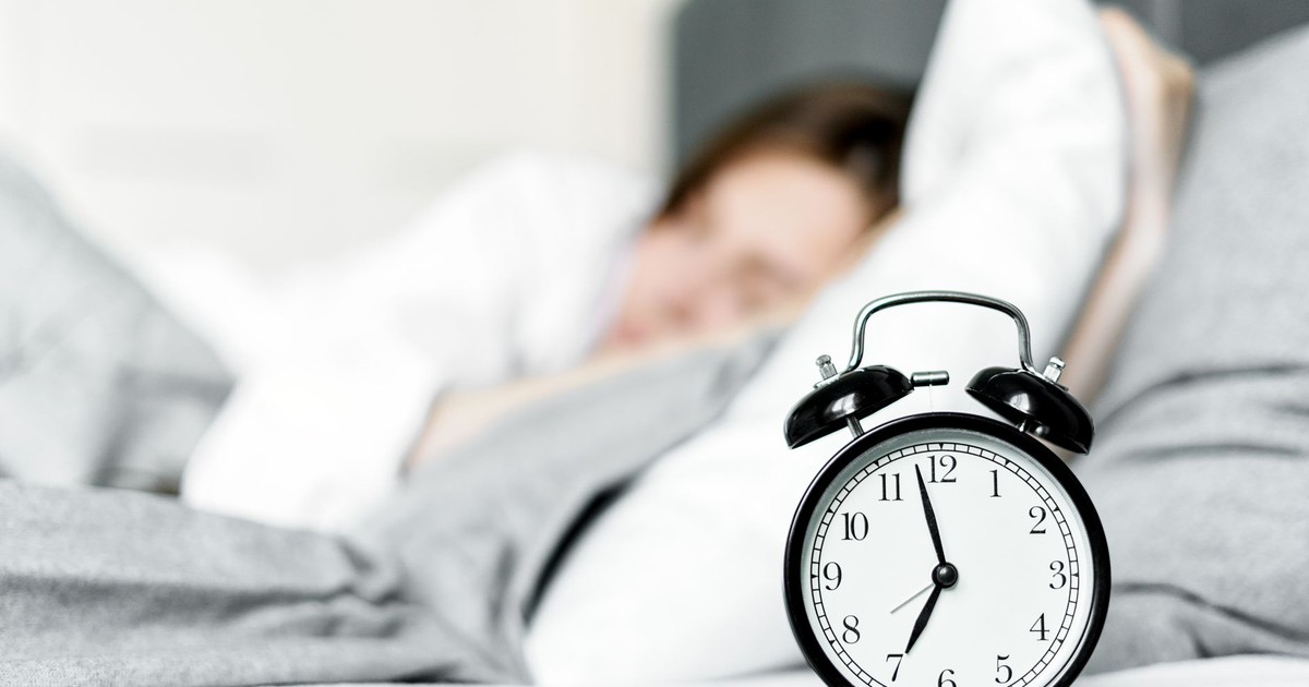 A retro alarm clock sits by a sleeping woman in a cozy bedroom setting.