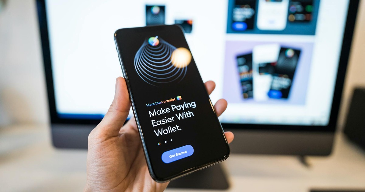 Hand holding smartphone displaying digital wallet app interface, blurred monitor in background.