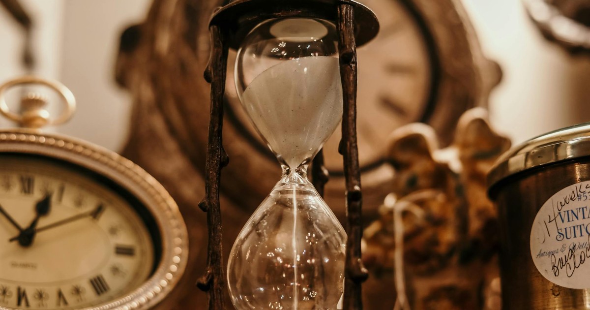 Close-up of a vintage hourglass surrounded by antique clocks in warm lighting.