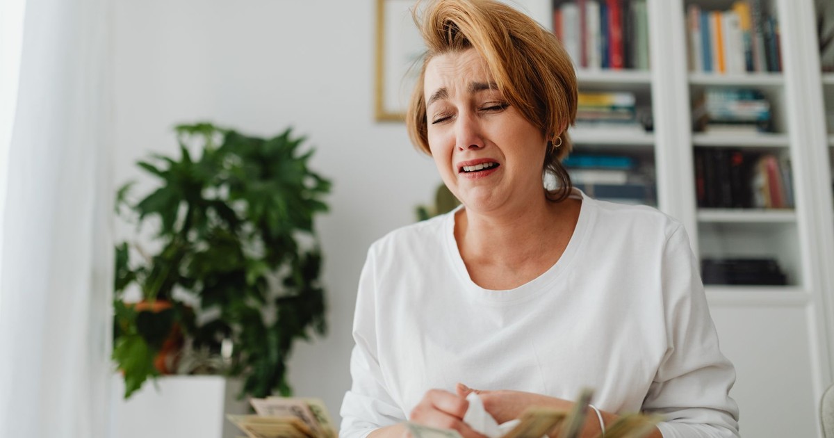 A distressed woman counts cash at a desk with a pained expression, highlighting financial strain.
