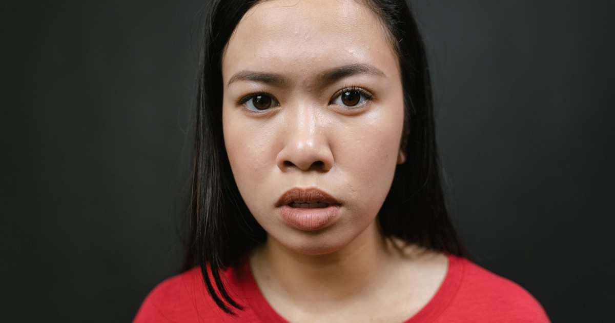 A close-up image of a woman in a red shirt against a gray background with a serious expression.
