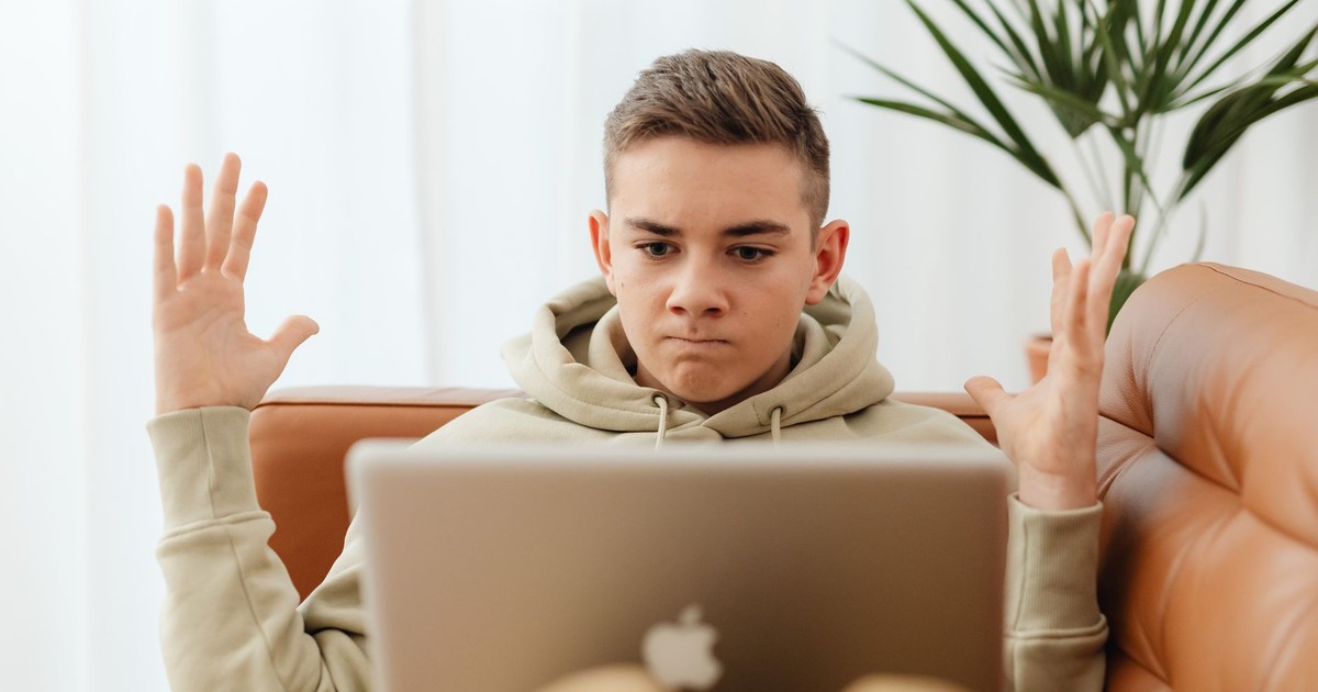 Teen sitting on couch with laptop, appearing frustrated, indoors.