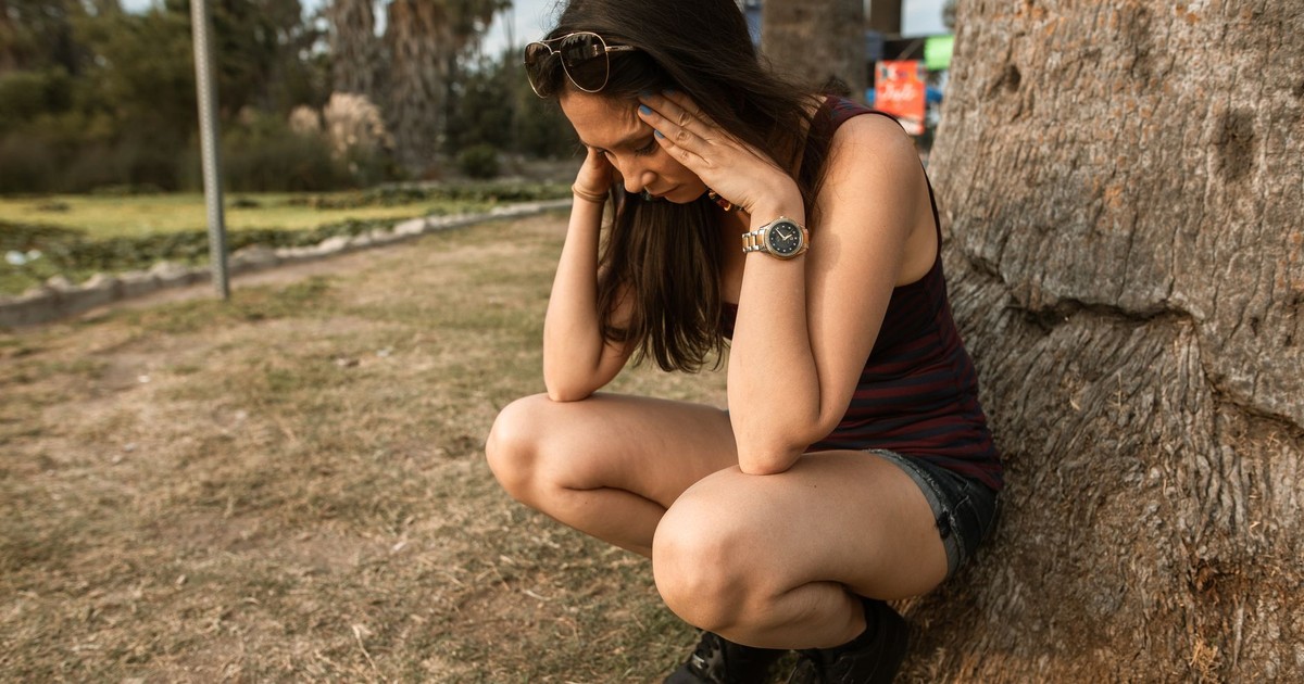 A woman kneels by a tree holding her head, expressing stress or anxiety in an outdoor park.