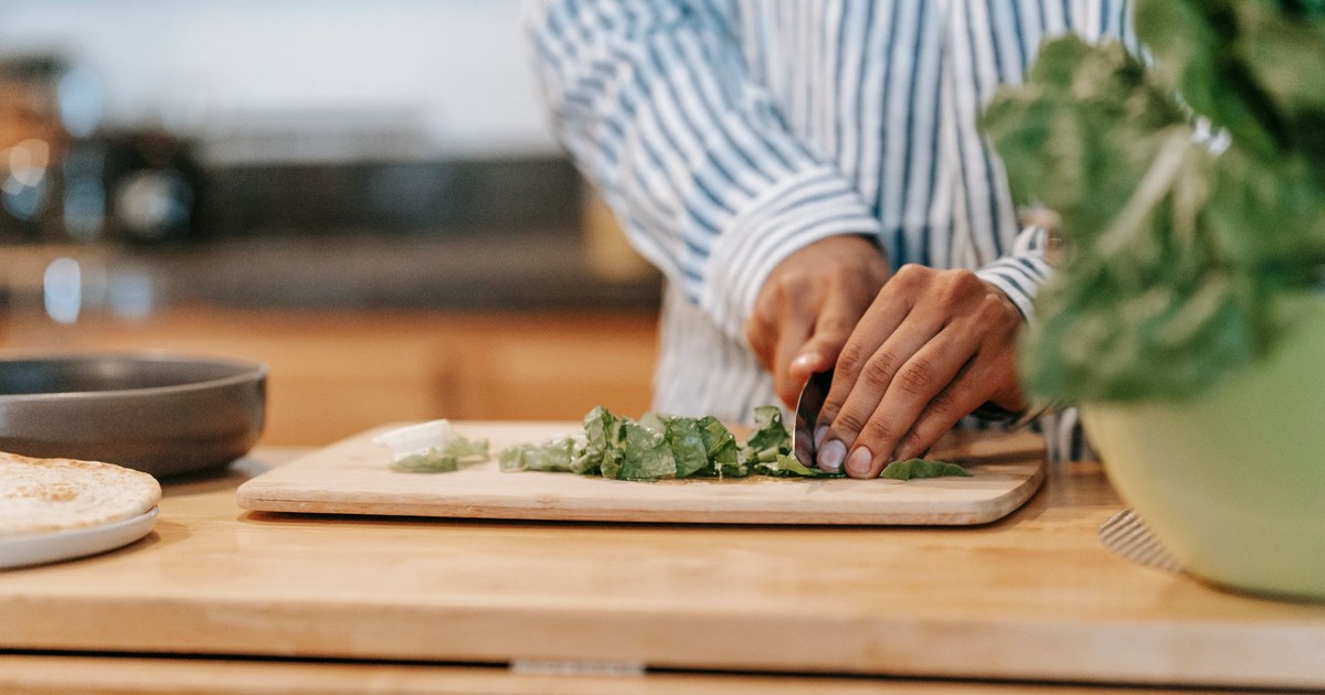Crop anonymous ethnic male in striped shirt cutting fresh spinach on chopping board while cooking at home