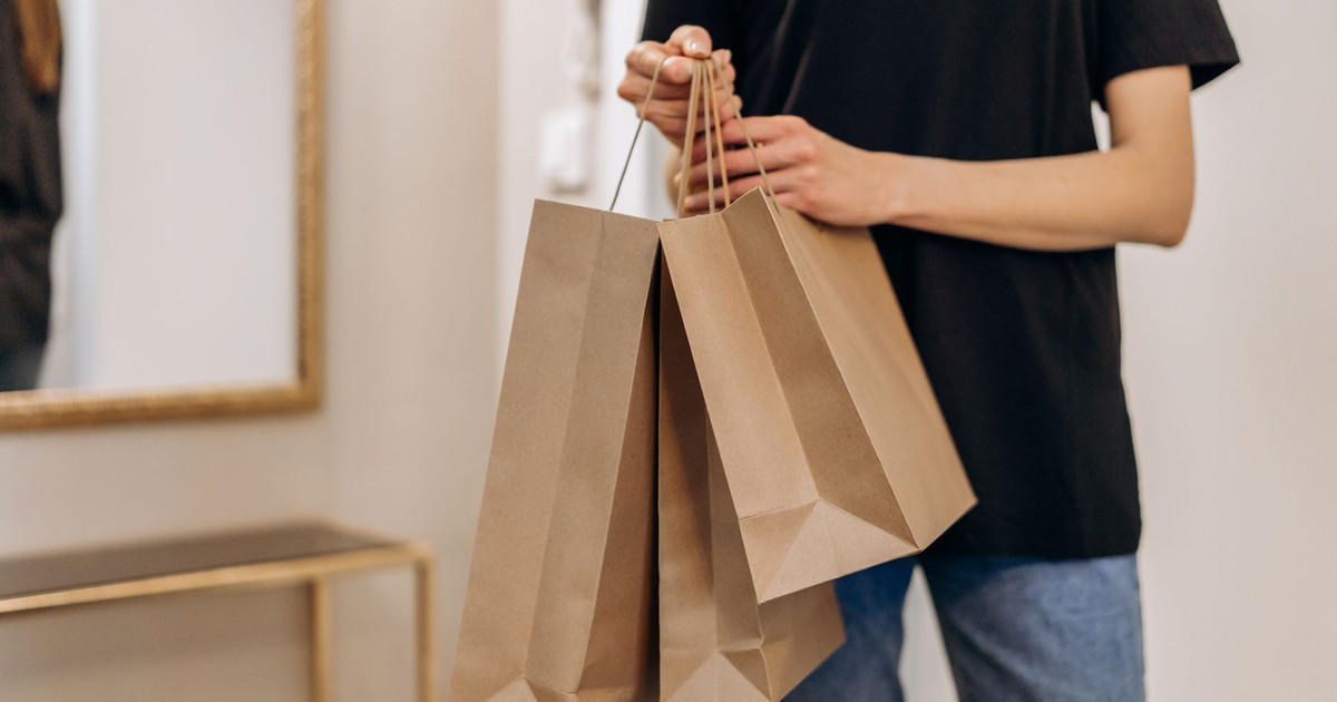 A person standing indoors holding three brown paper shopping bags in one hand, wearing casual clothing.
