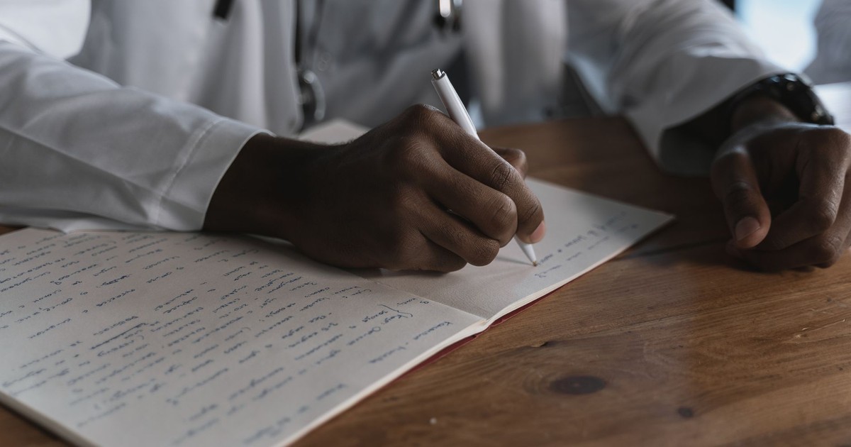 Close-up of a doctor writing notes with a pen in a medical notebook on a wooden desk.