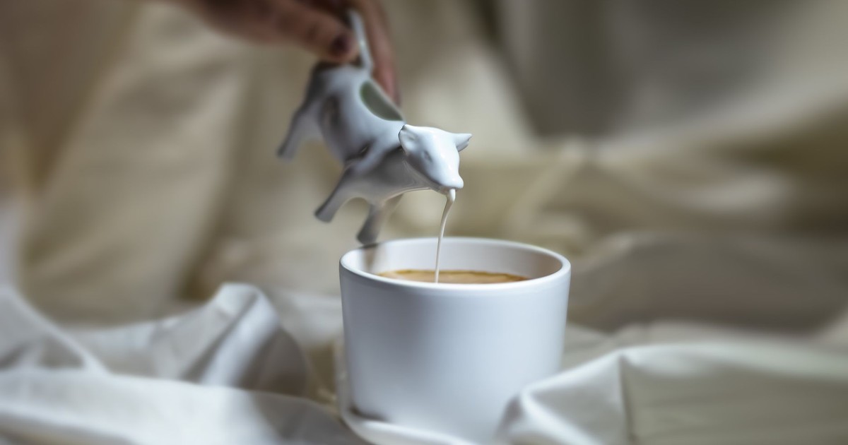 A hand pours milk from a cow-shaped creamer into a cup of coffee on a soft fabric background.