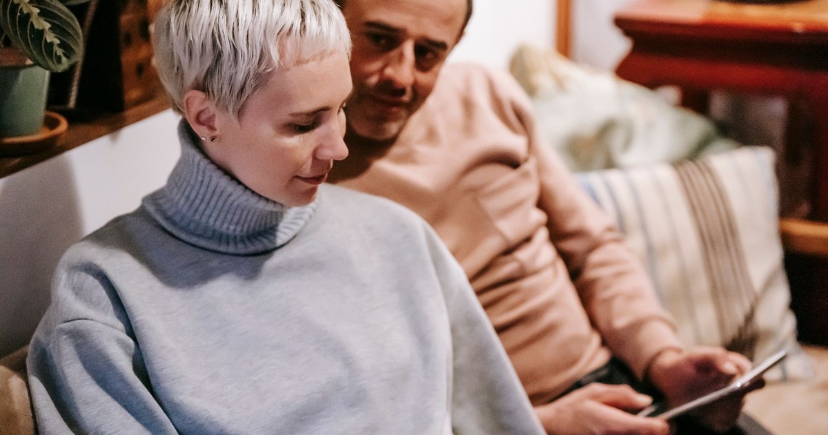 Calm female with short hair in stylish sweater sitting on sofa near ethnic adult husband using tablet during weekend at home