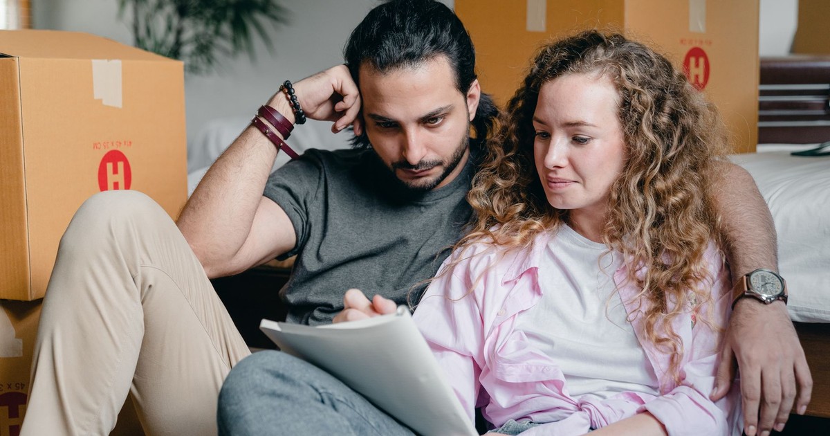 Pensive male and female in casual clothes sitting together among boxes and writing notes in notebook while leaning on bed