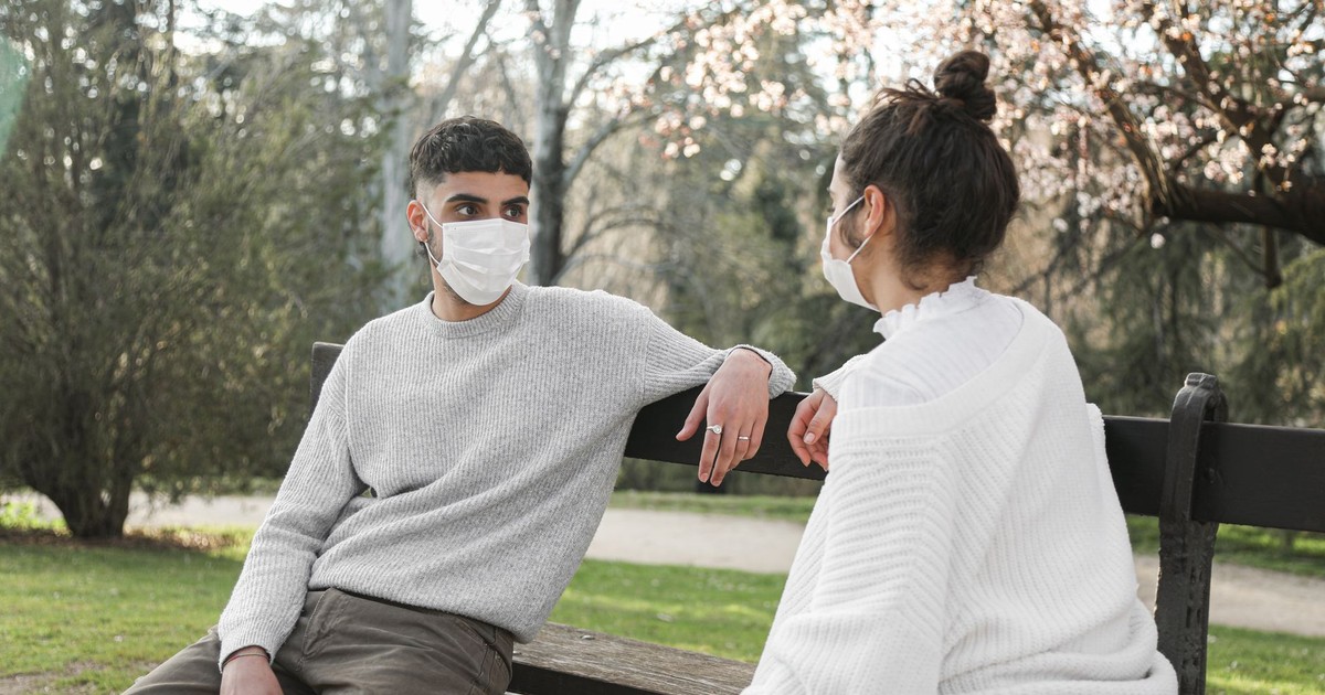 Young couple wearing face masks seated on a bench in a park engaging in conversation.