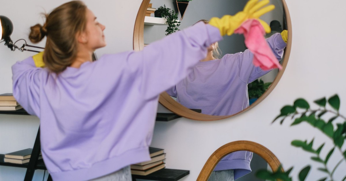 Back view of young female in casual clothes and yellow latex gloves dancing while cleaning light living room in daytime