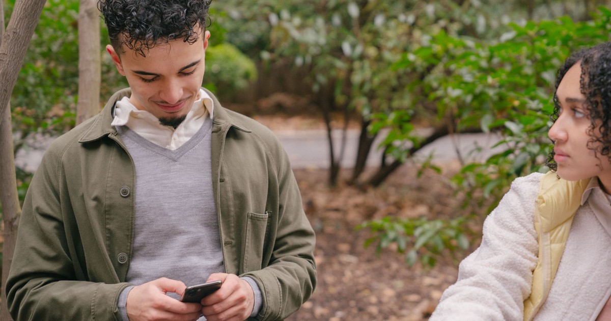 Hispanic happy boyfriend surfing internet on mobile phone while focused crop girlfriend watching in park with green plants