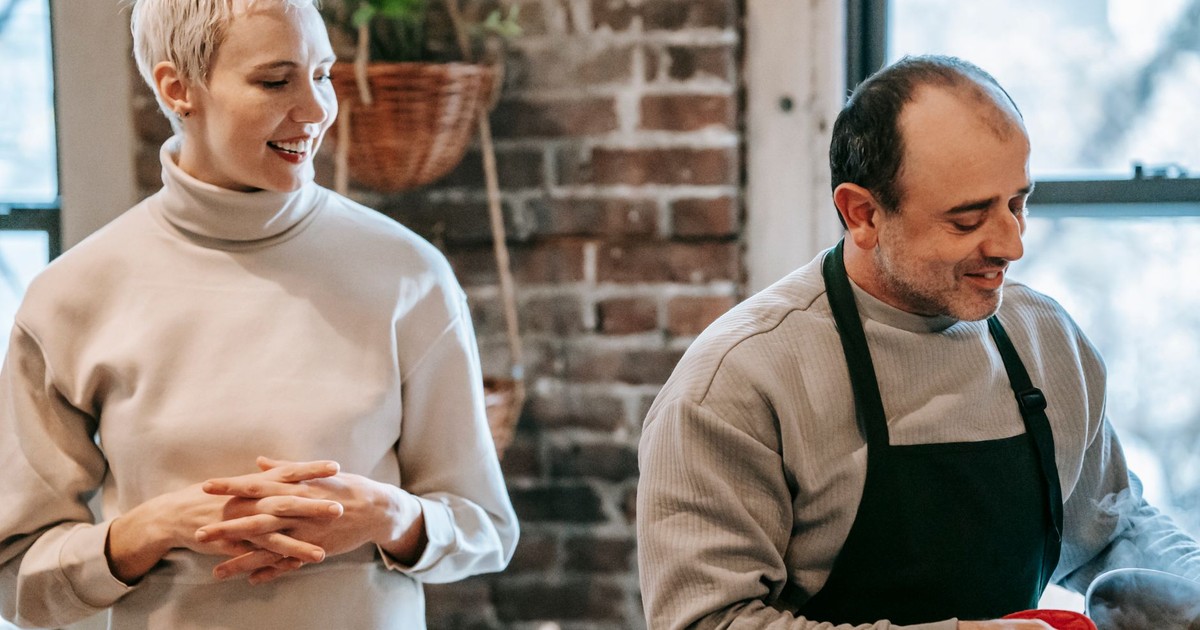 Positive adult woman in casual clothes smiling and looking at ethnic husband in apron preparing delicious dinner in kitchen