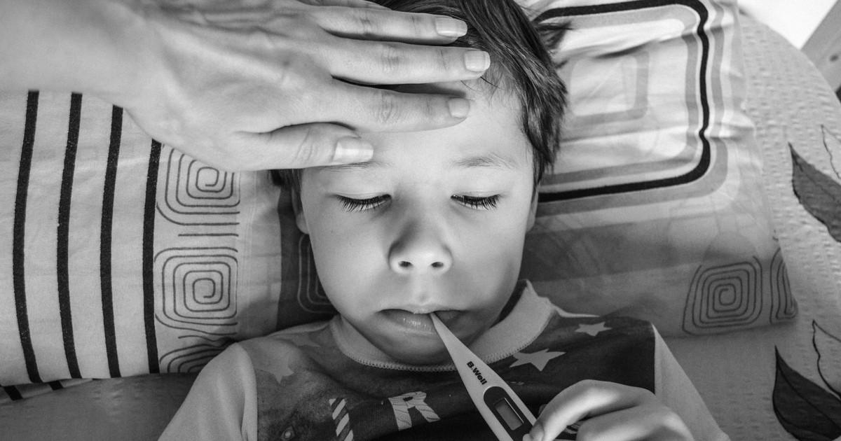 Black and white from above of unrecognizable person touching forehead of unemotional child in casual clothing holding electronic thermometer in mouth