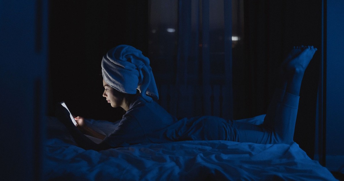 A woman lying in bed with a towel on her head, using a mobile phone at night.