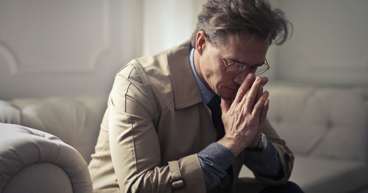 Side view of upset male entrepreneur in formal wear sitting on couch in modern living room and contemplating about financial problems while leaning on hands and looking down