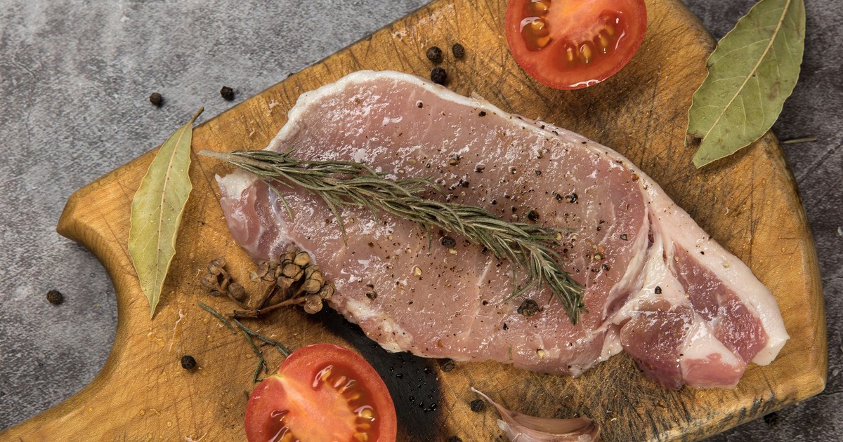 Raw pork chop with seasoning, herbs, and tomatoes on a wooden board.