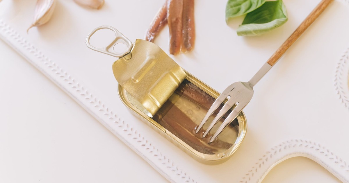 Top view of an open can of anchovies with fork, basil, and garlic on white tray.