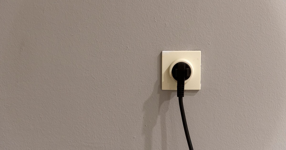 A black plug inserted into a white wall socket on a plain grey wall.