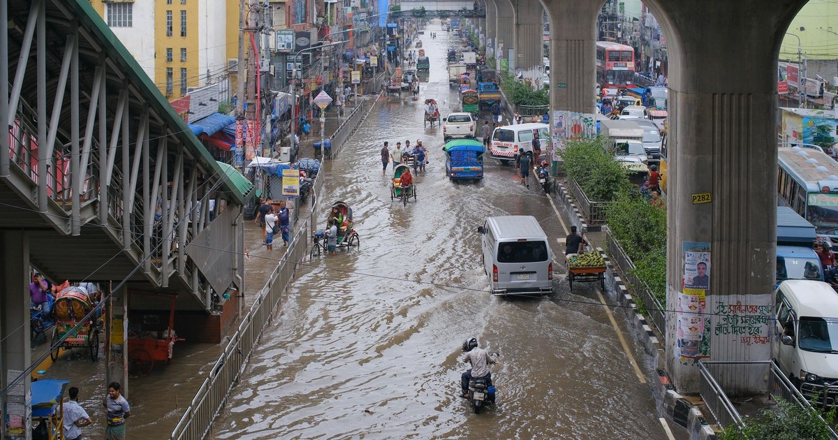 Urban street flooded with vehicles and pedestrians navigating water under elevated metro tracks.