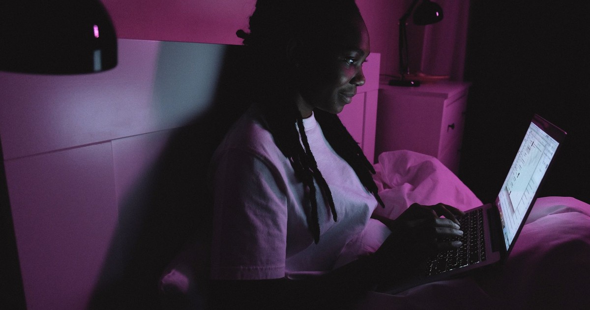 An adult woman using a laptop in bed under purple lighting, representing modern remote work at home.