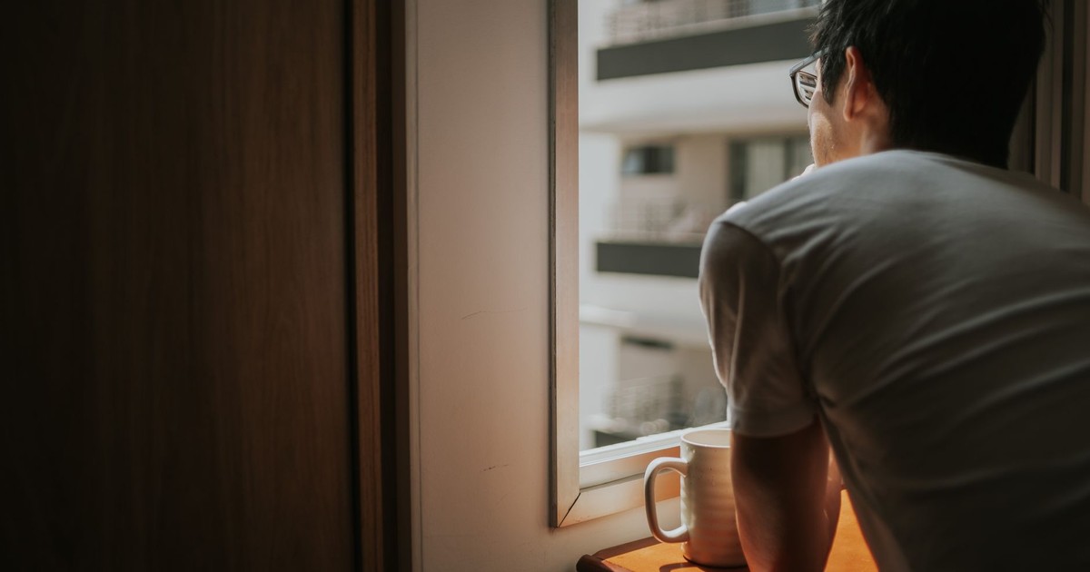 A man thoughtfully looks out a window with city buildings in view, holding a mug.