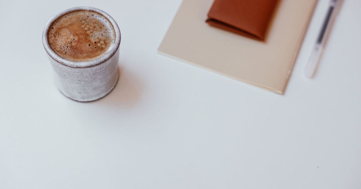 A minimalist setup with a cup of coffee, a notebook, and a pen on a white tabletop.