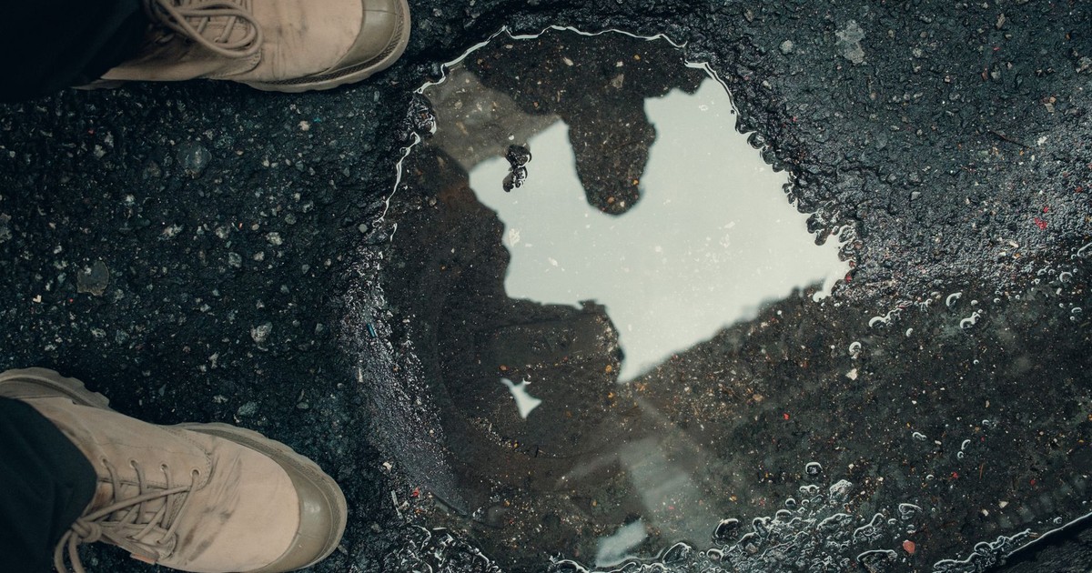 Close-up of shoes and a puddle on an asphalt surface reflecting people.