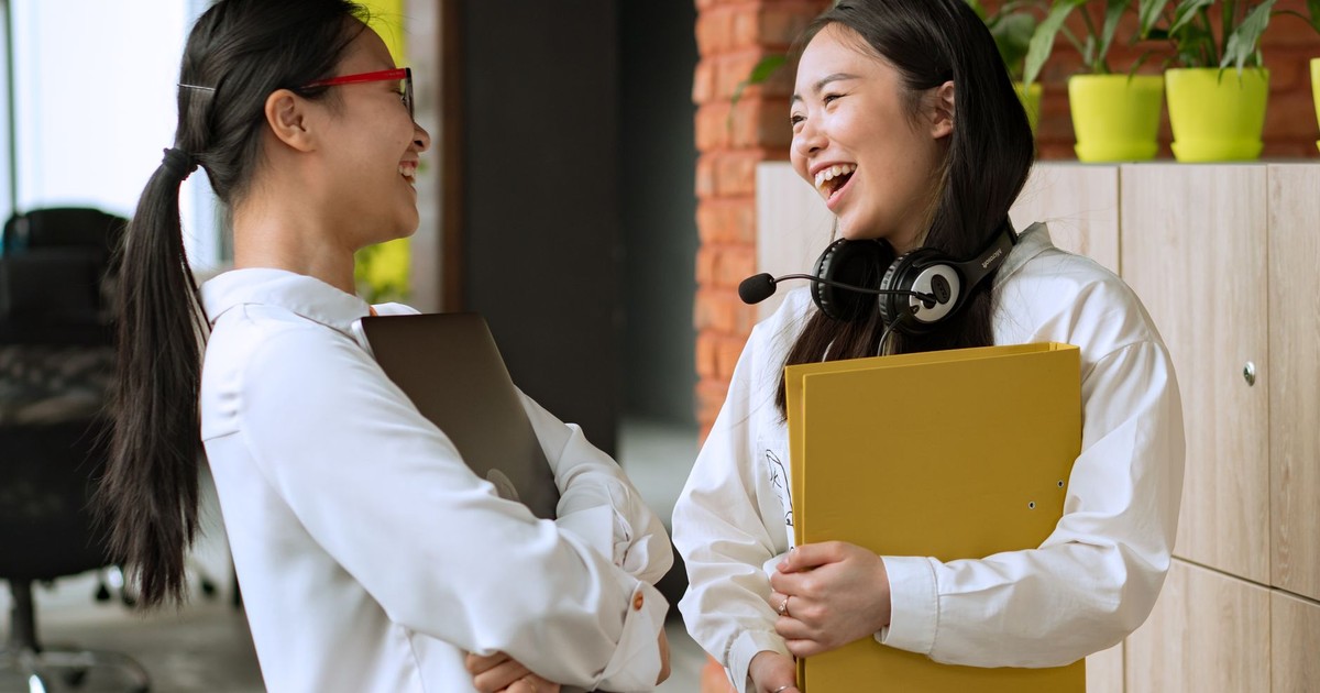 Two women sharing a light moment in a modern office, holding folders and wearing headphones.
