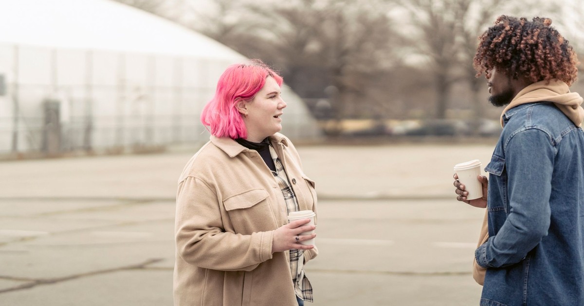 Plus size woman with pink hair enjoying coffee to go and talking with African American man while standing on city street on autumn day
