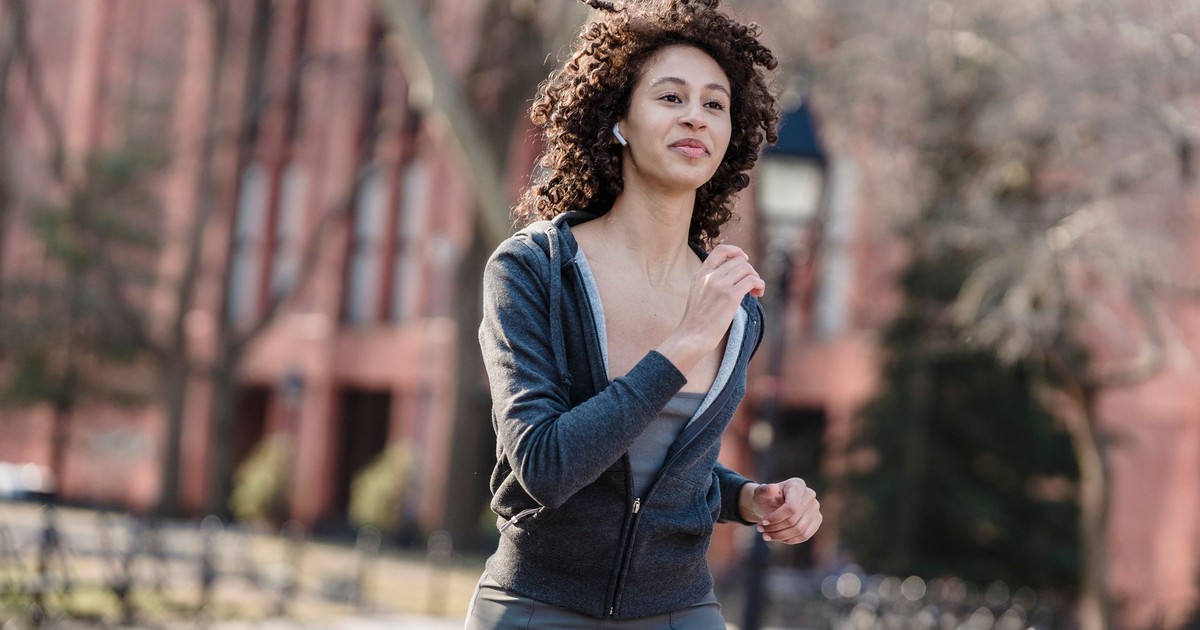 Positive curly haired female in activewear listening to music in wireless earbuds while jogging in park