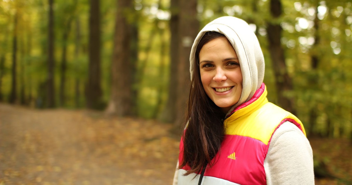A brunette woman in a hoodie and vest smiles while hiking in a forest in Slovakia.